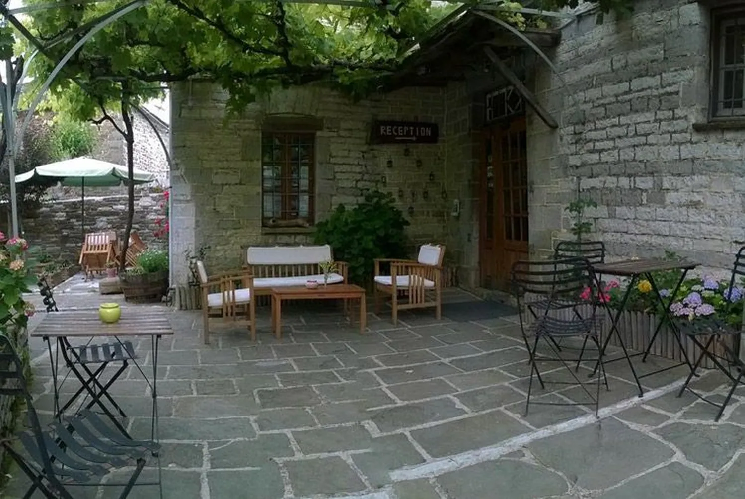 Πλακόστρωτη αυλή με κληματαριά, καρέκλες και τραπεζάκια μπροστά από πέτρινο σπίτι